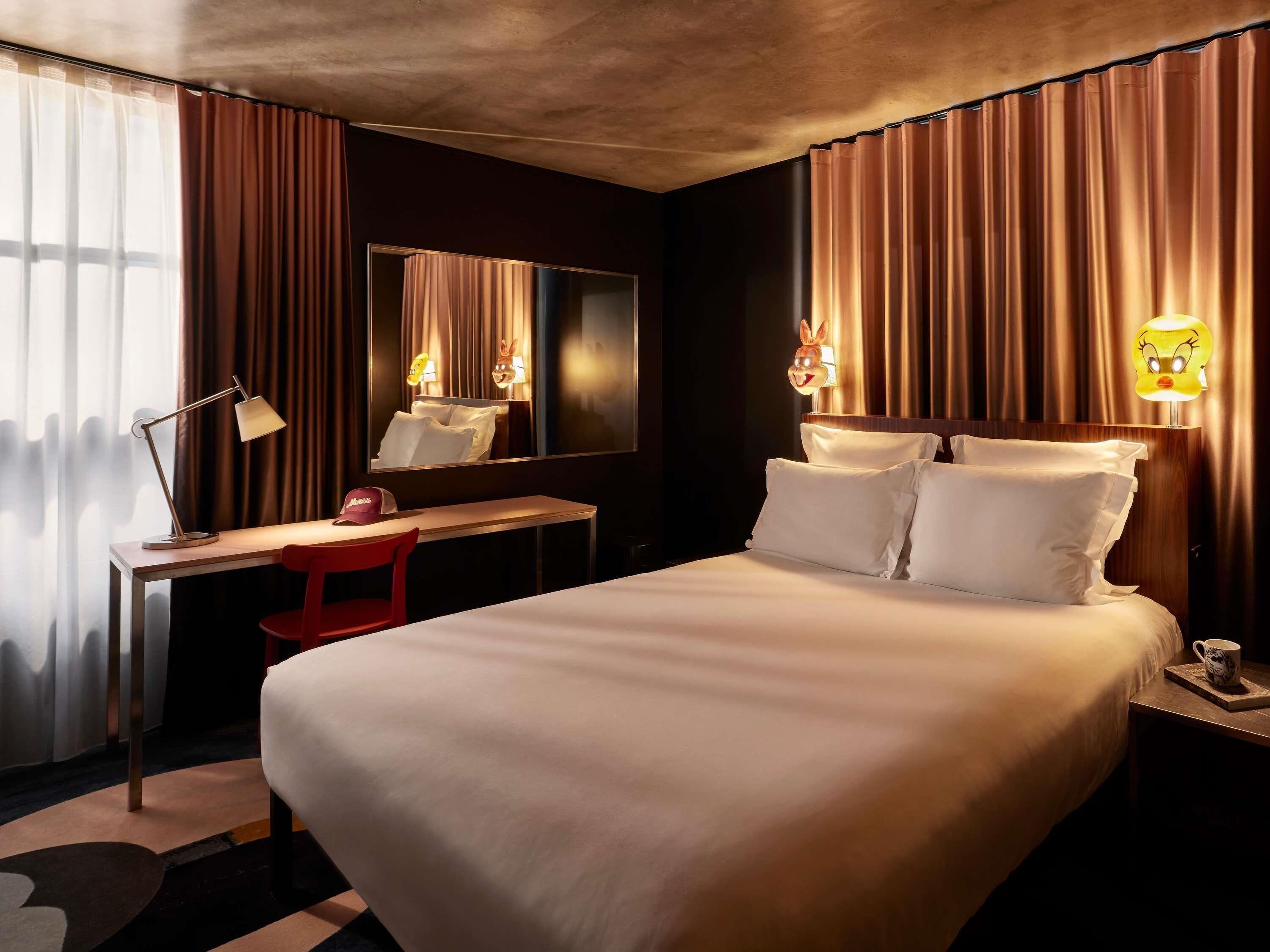 A hotel room with a large bed, a desk with a chair, a mirror, and two wall-mounted lamps.