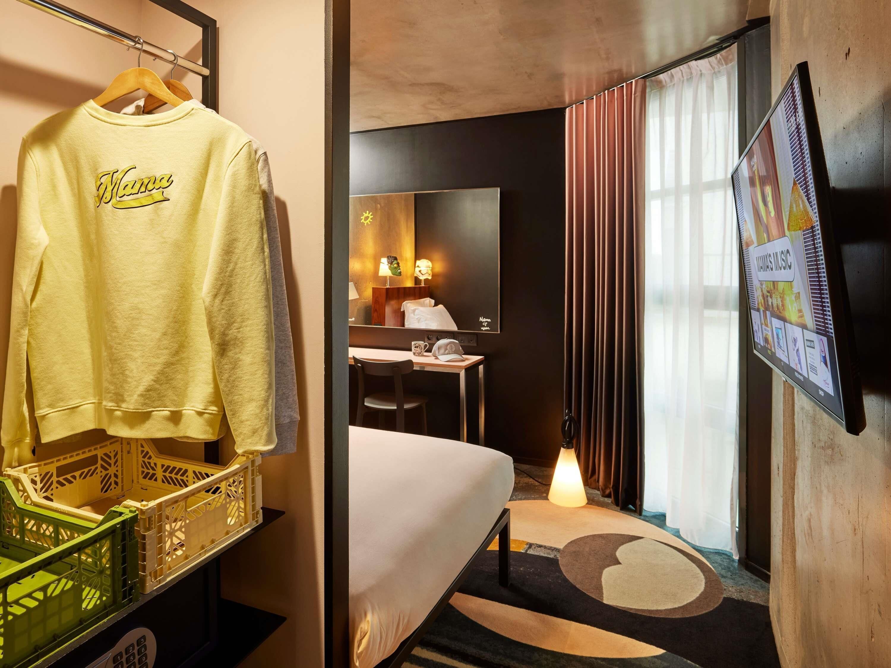 A hotel room with a bed, a desk, a chair, a TV, and a wall-mounted screen displaying music.
