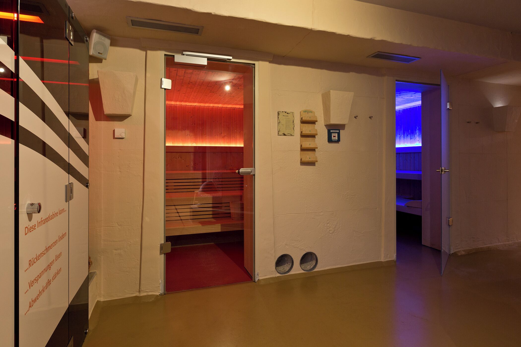 Un pasillo con una habitación iluminada con luz roja, una puerta con un letrero y una rejilla en la pared.