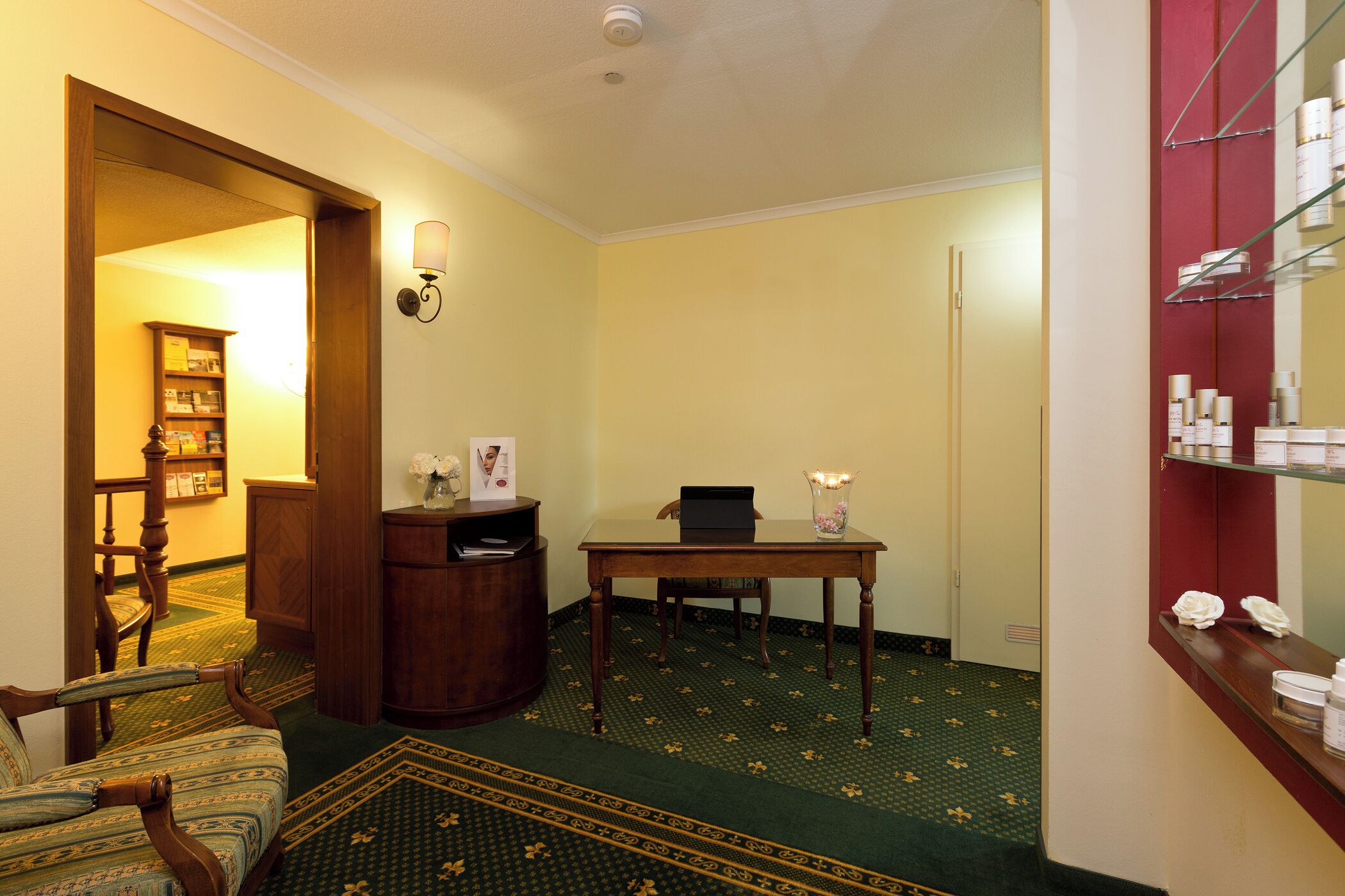 A room with a green patterned carpet, a wooden chair, a small table with a vase, a wooden cabinet, and a mirror with shelves.