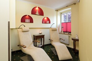 A room with two reclining chairs, a small table with a lamp, and a window with blinds.