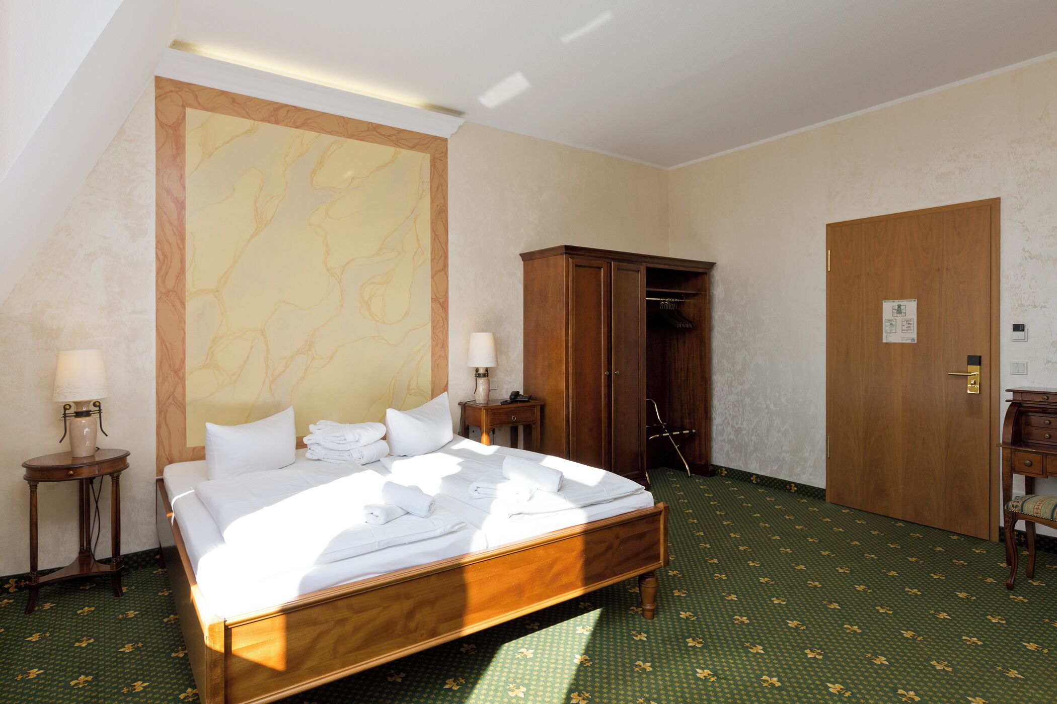 Una habitación de hotel con una cama grande, un armario, una mesita de noche con lámpara y una alfombra verde con estampado.