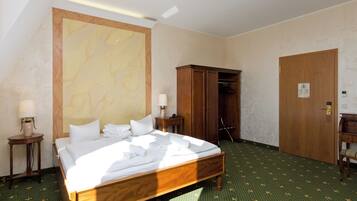 Una habitación de hotel con una cama grande, un armario, una mesita de noche con lámpara y una alfombra verde con estampado.