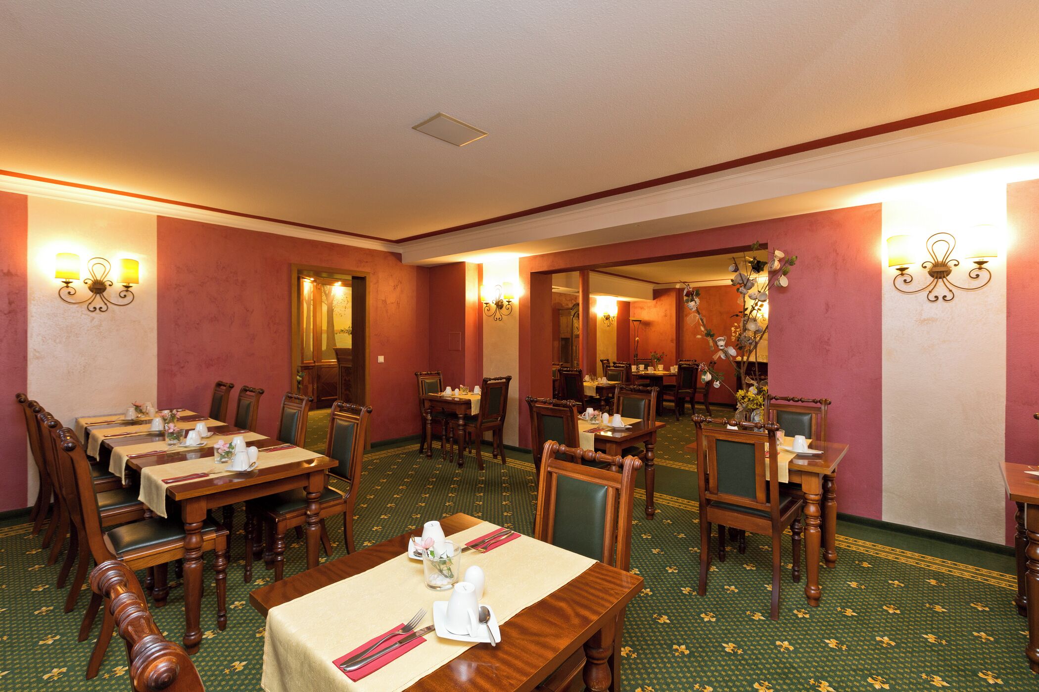 A hotel dining area with multiple tables and chairs, a patterned carpet, and wall-mounted lighting fixtures.