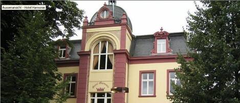 A two-story building with a yellow facade and red trim, featuring a central entrance with a canopy and a sign that reads "Hotel Harmonie."
