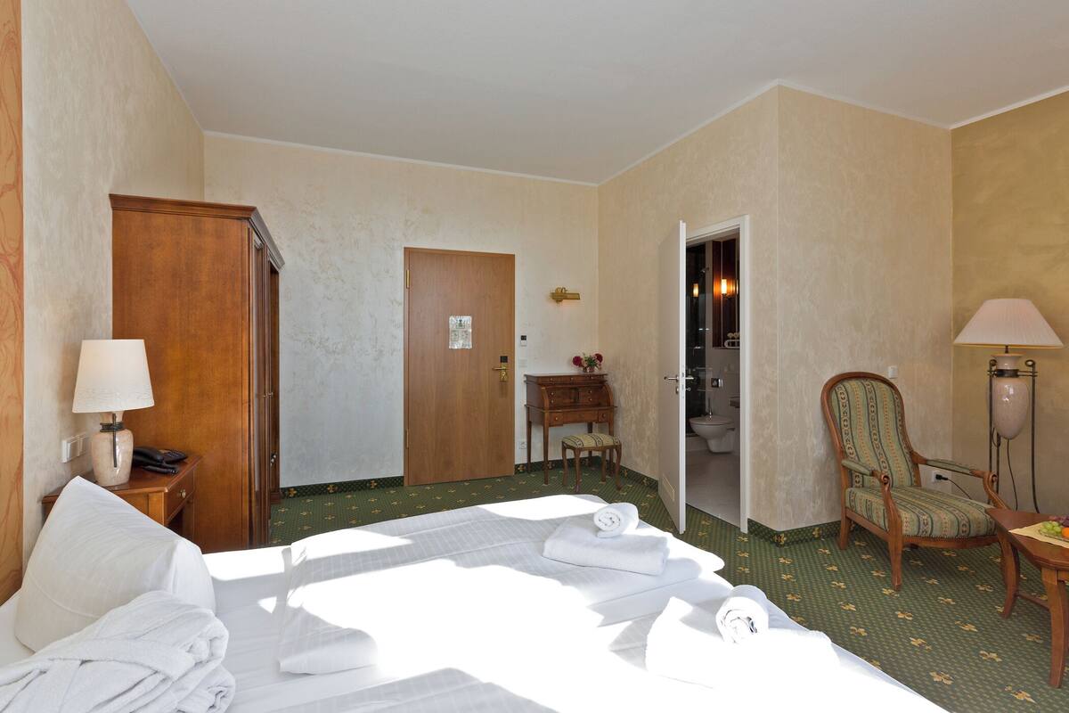 Ein Hotelzimmer mit einem Bett, einem Holzschrank, einem Schreibtisch mit Stuhl, einer Lampe und einem durch eine offene Tür einsehbarer Badezimmer.