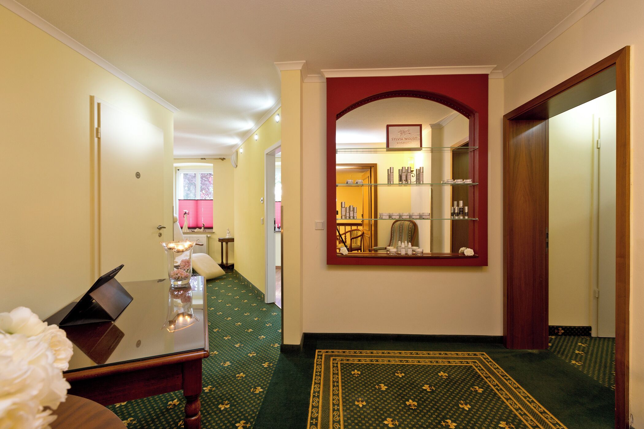 A hotel hallway with a green carpet, a mirror with a red arch, and a shelf with toiletries.