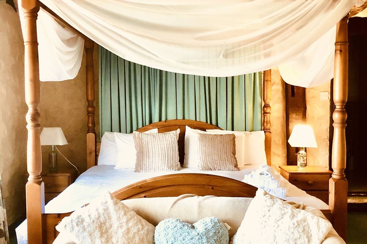 A four-poster bed with a canopy, multiple pillows, a heart-shaped cushion, and bedside lamps.