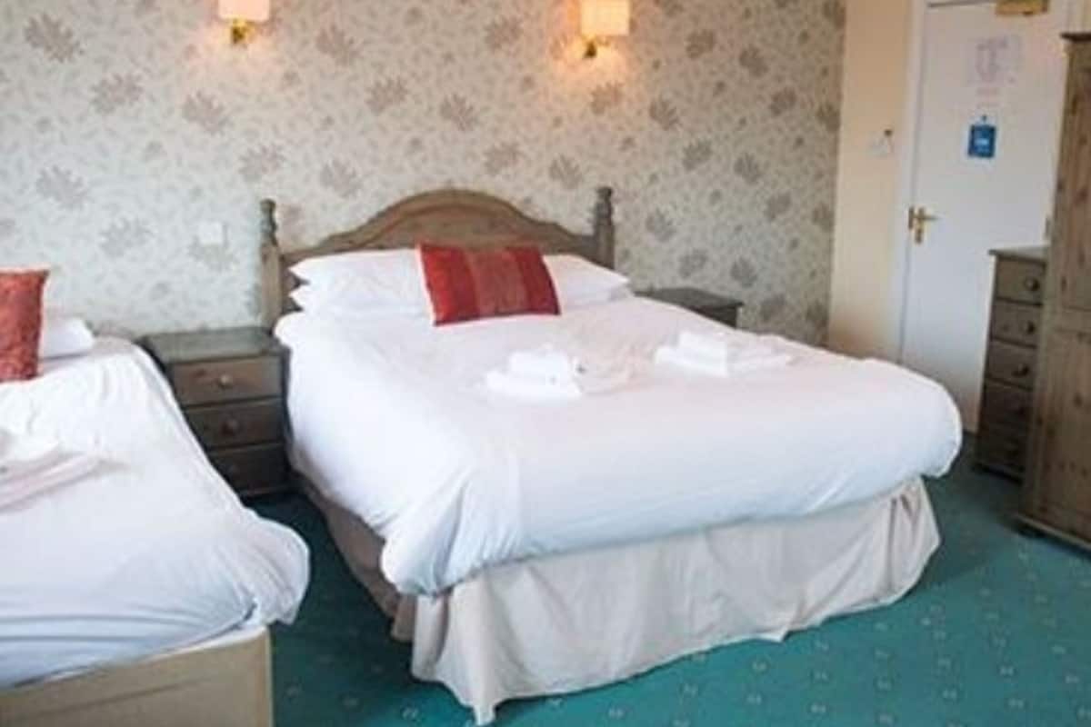 A hotel room with two beds, a wooden headboard, a nightstand, a dresser, and a patterned wallpaper.