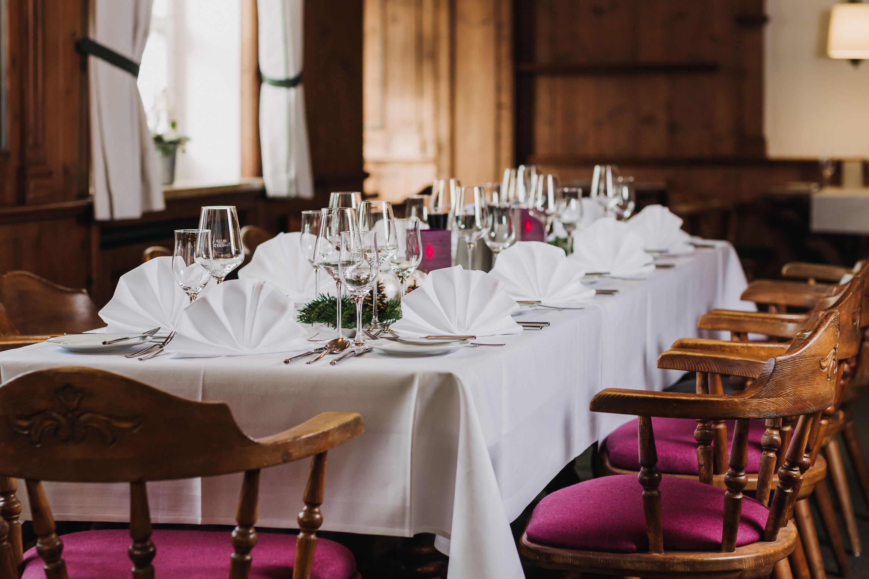 A long dining table set with white tablecloths, glassware, and wooden chairs with purple cushions.