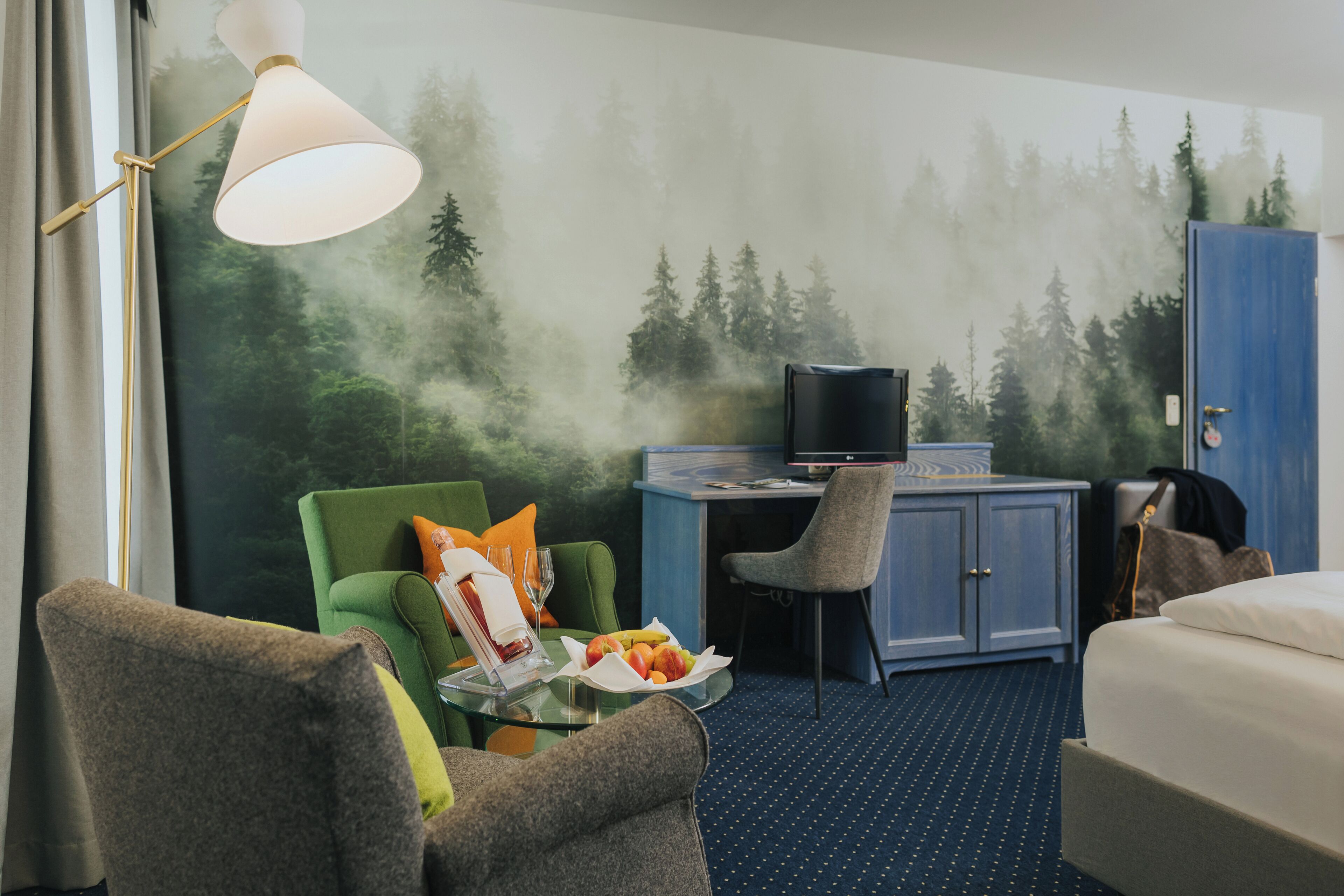 A hotel room with a bed, a green armchair, a grey armchair, a small table with a fruit plate, a lamp, a TV, and a wardrobe.