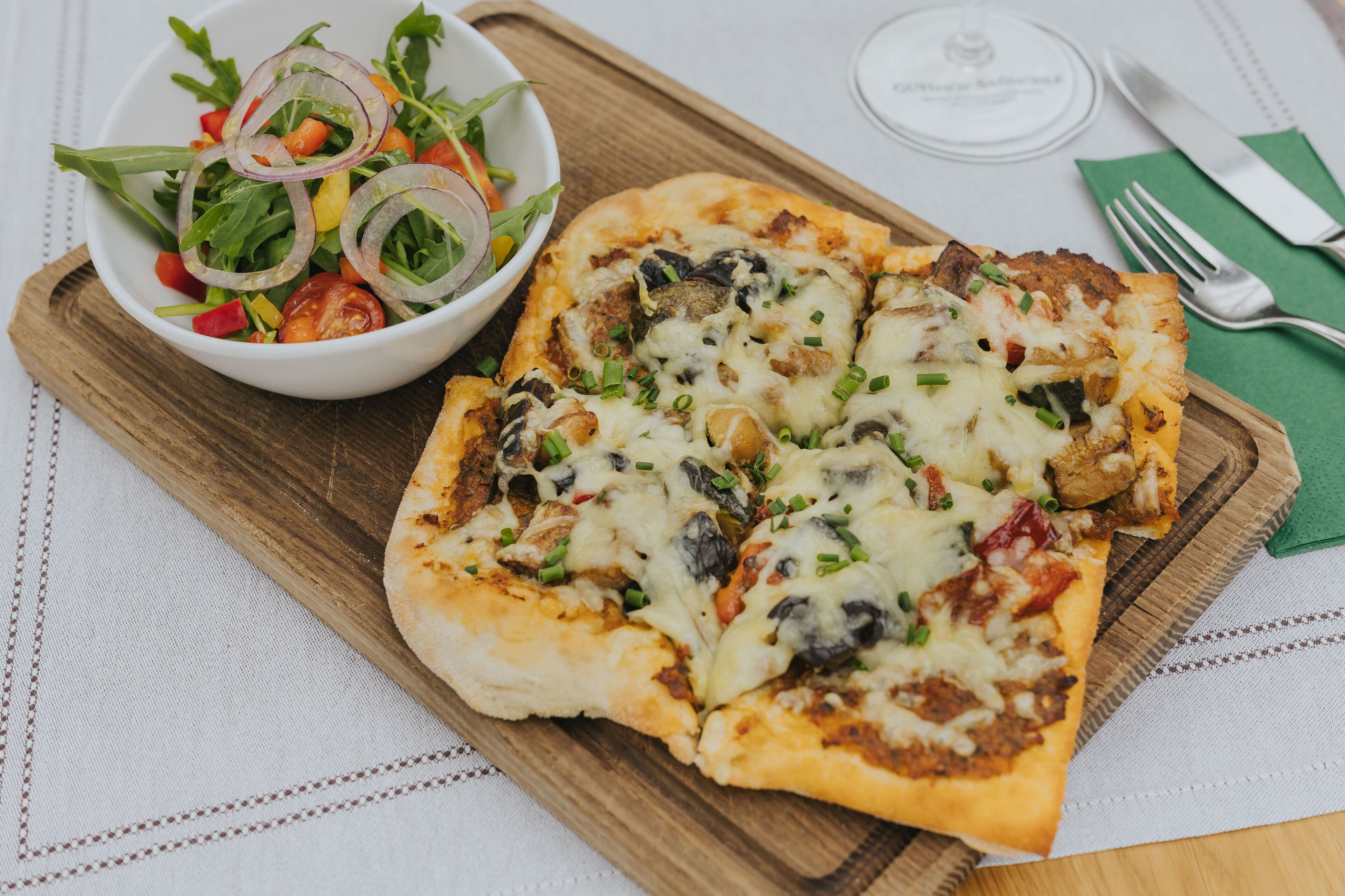 A pizza with toppings on a wooden board, a salad with mixed greens and sliced onions, and a green napkin with cutlery on a tablecloth.