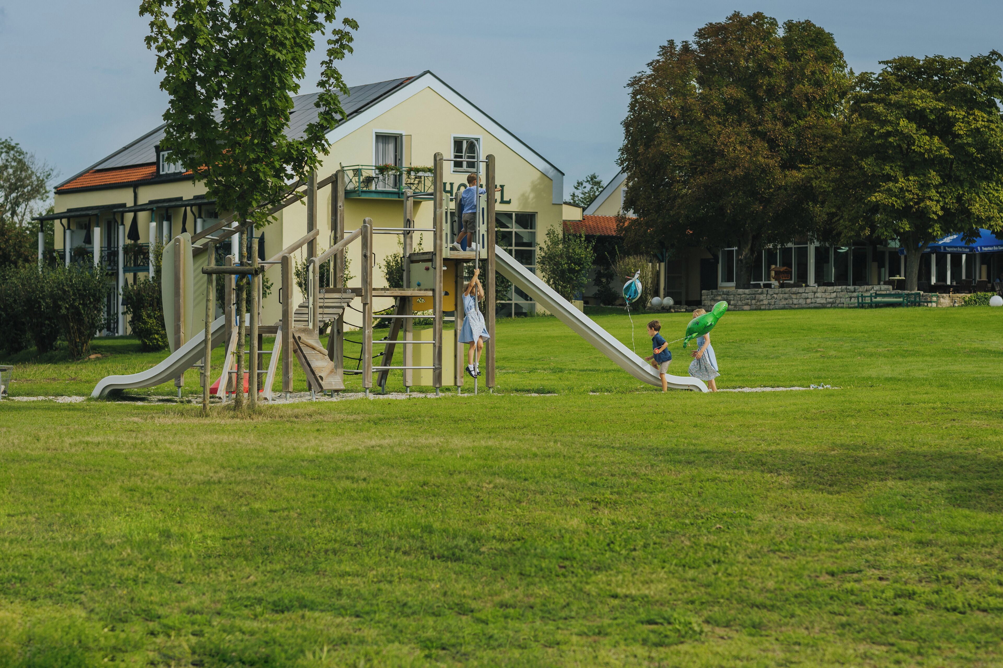 A playground with a slide and climbing structures, set in a grassy field with a building in the background.