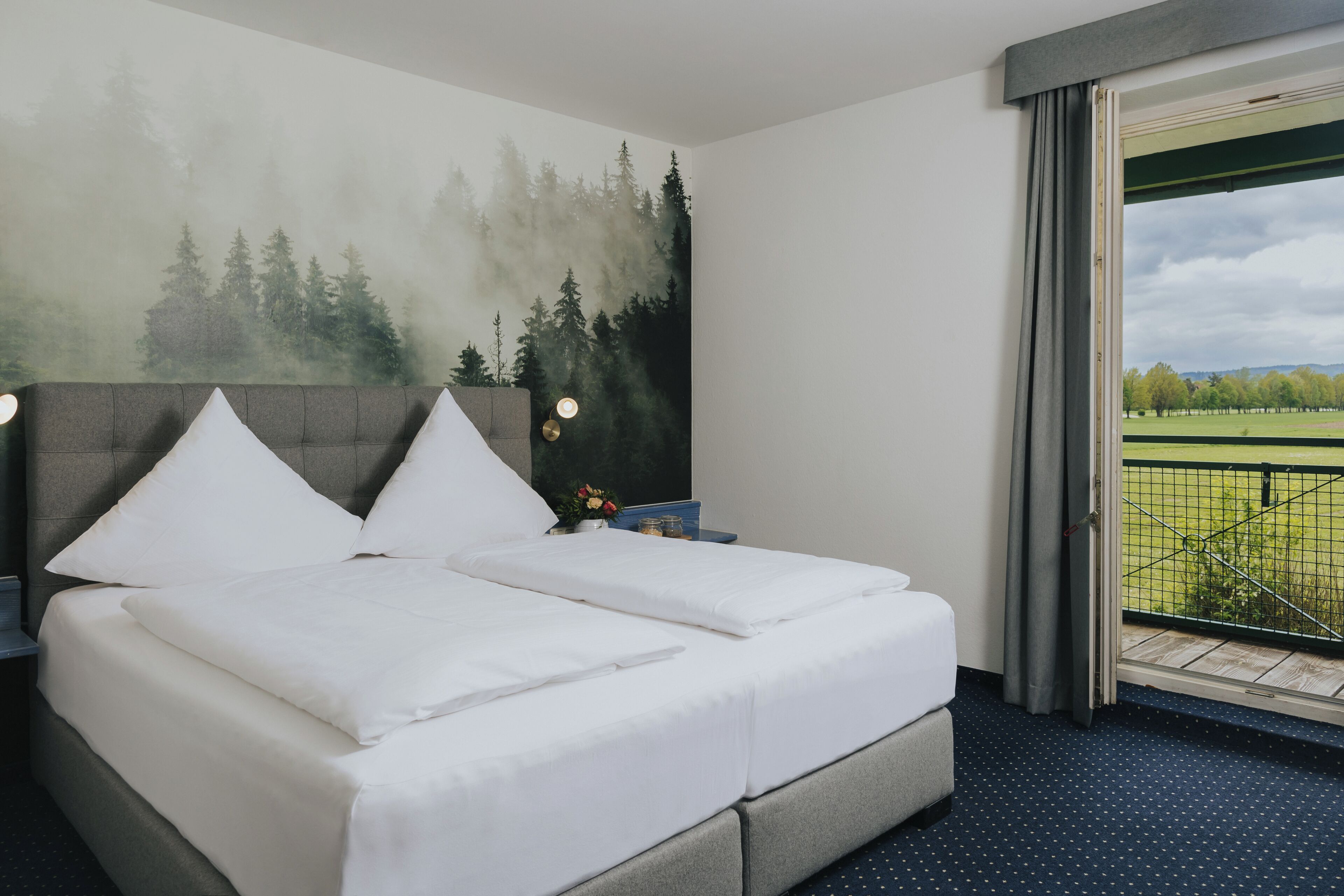 Una habitación de hotel con cama, mesita de noche, ventana con vistas a un parque y un cuadro en la pared.