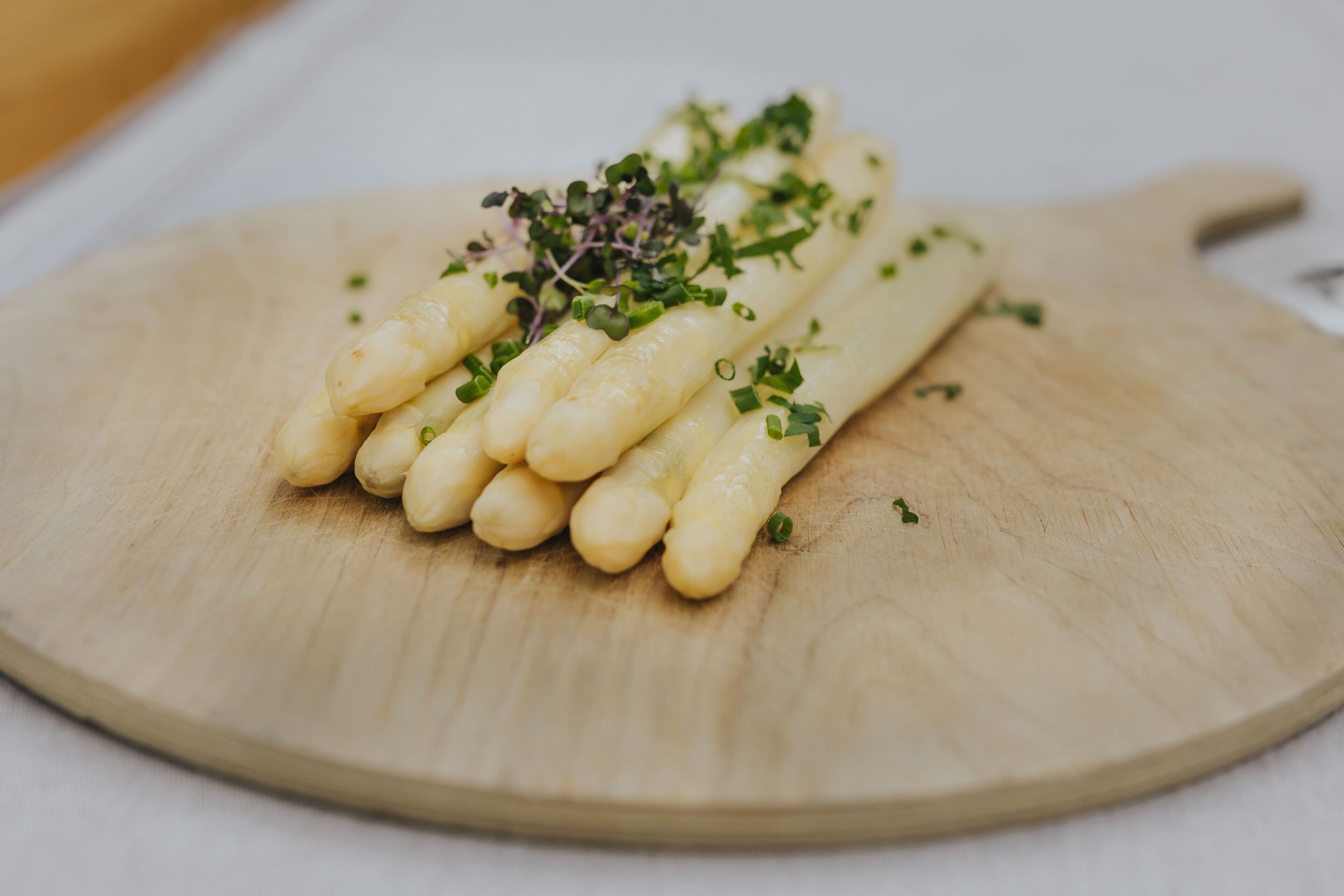 A wooden cutting board with a bunch of white asparagus spears garnished with green herbs.