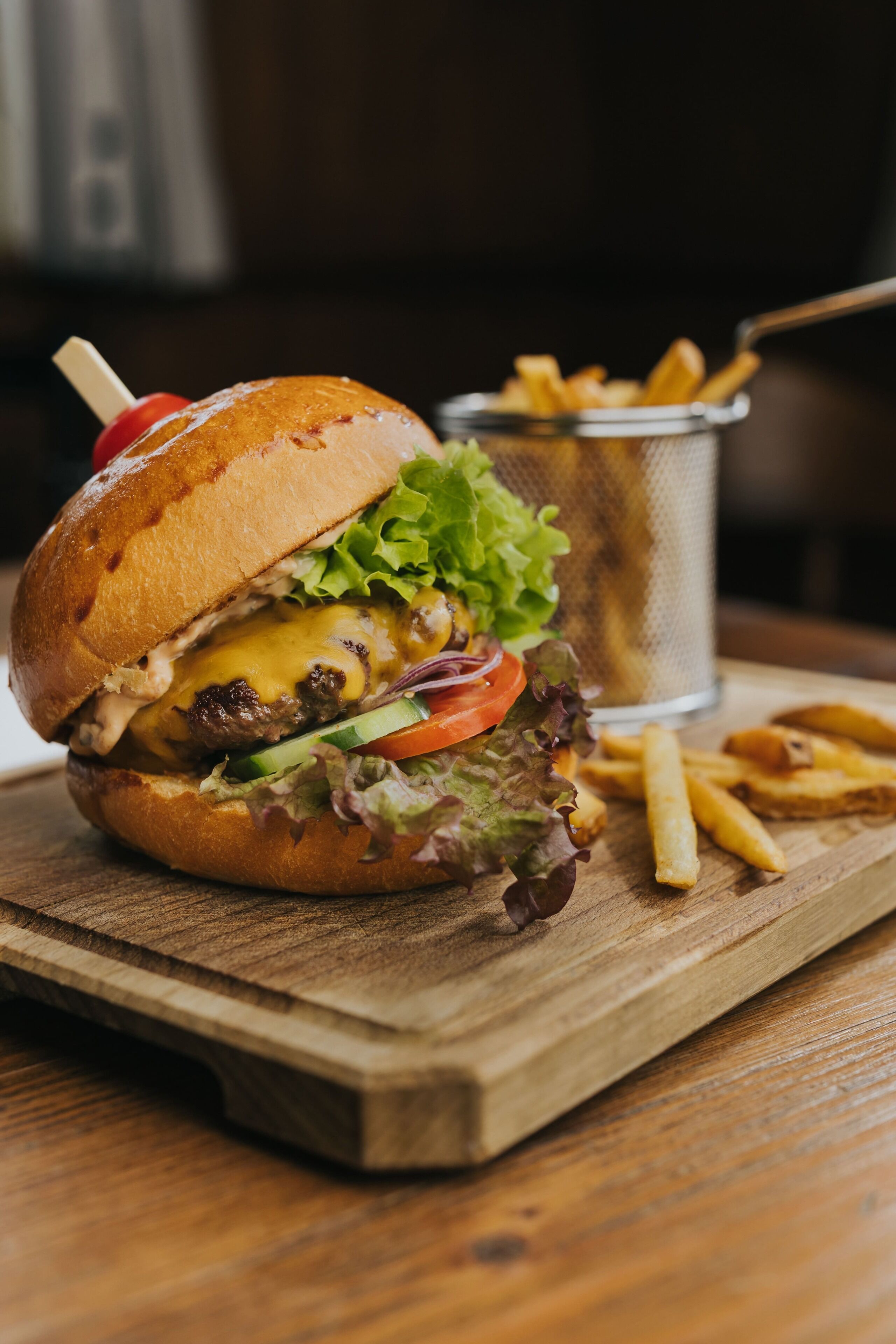 A burger with lettuce, tomato, and cheese on a bun, accompanied by a side of fries on a wooden board.