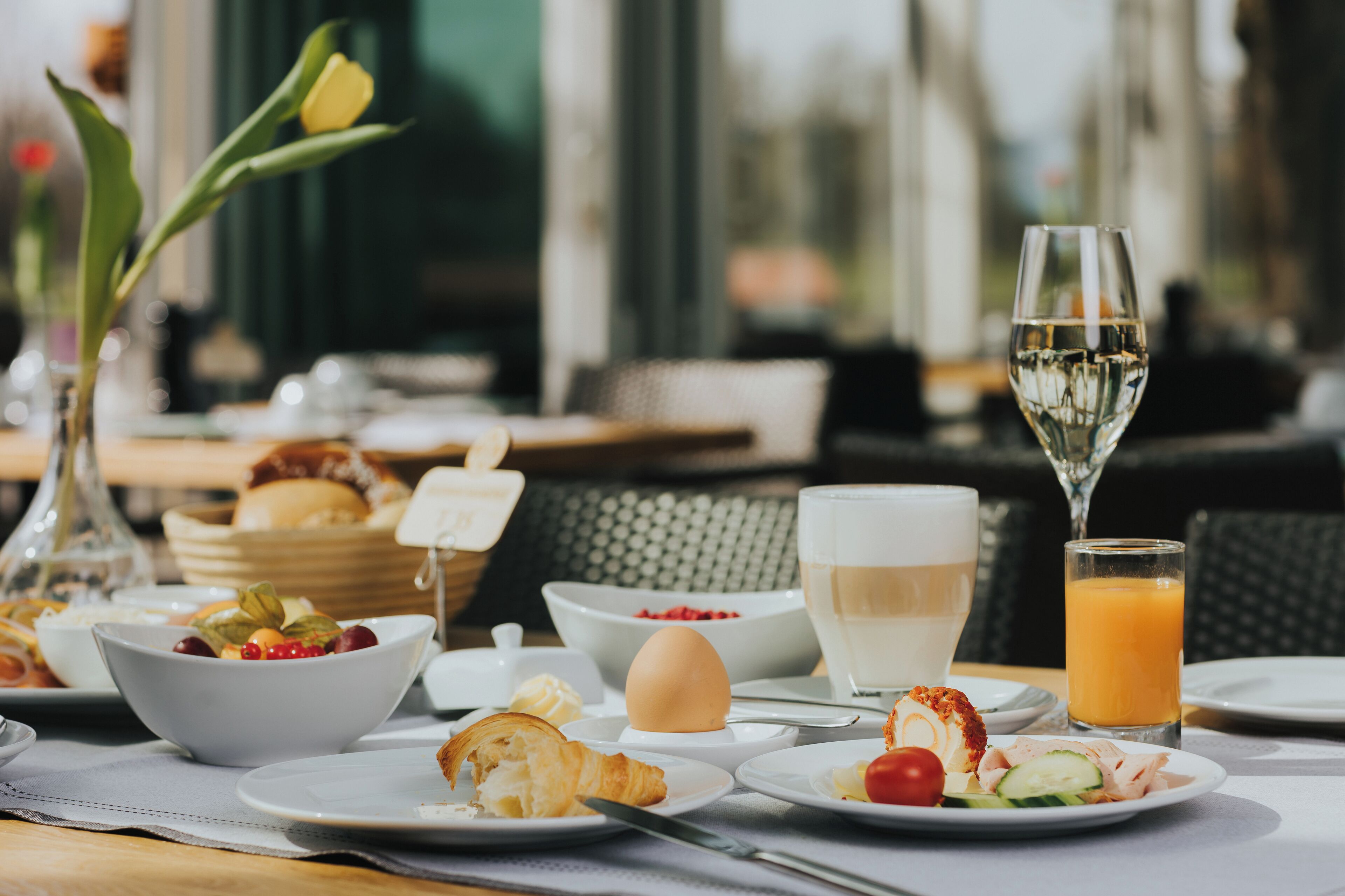 A breakfast table with various dishes, including eggs, toast, and fruit, along with a glass of orange juice and a glass of white wine.
