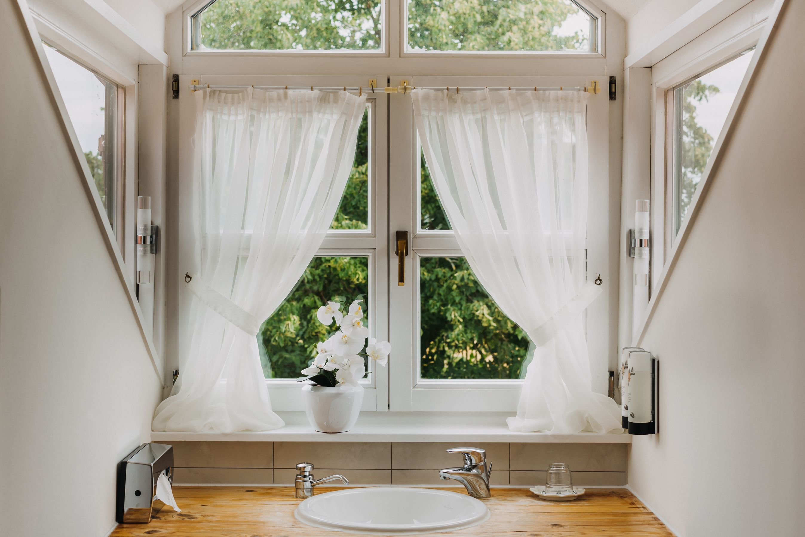A bathroom with a large window, white curtains, a white sink, and a potted orchid.