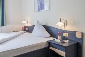 A hotel room with a bed, two bedside tables, a nightstand, a wall-mounted lamp, a framed picture, and a small potted plant.