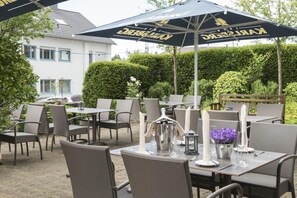 An outdoor dining area with tables and chairs, umbrellas, and a hedge in the background.