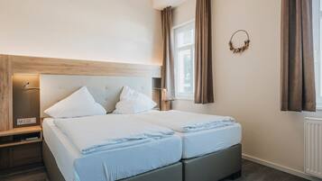 A modern hotel room with a large bed, a bedside table, a nightstand, a wall-mounted light, and a window with curtains.