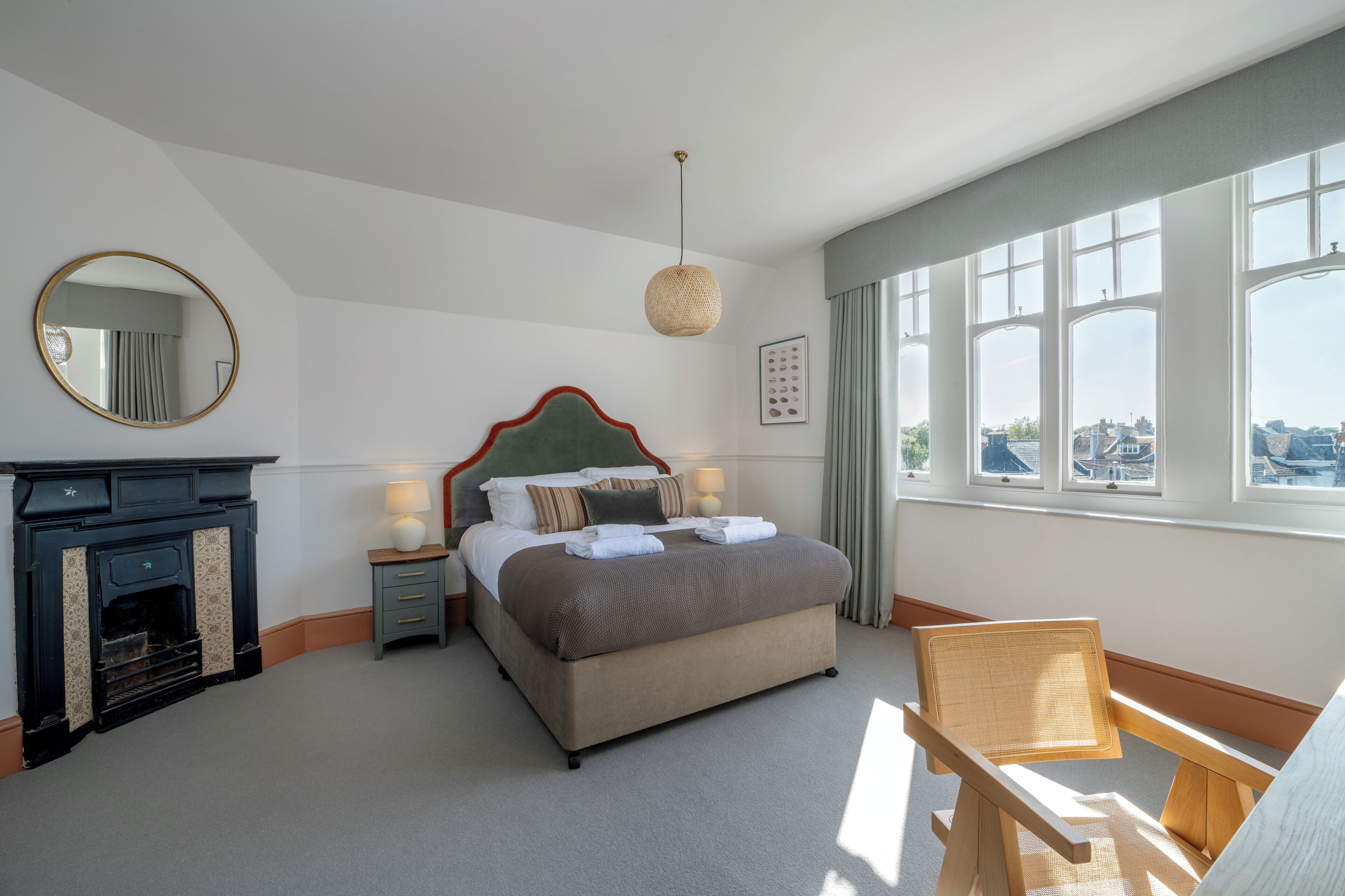 A neatly arranged bedroom with a bed, a small table with a lamp, a chair, a fireplace, and large windows offering a city view.