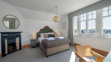 A neatly arranged bedroom with a bed, a small table with a lamp, a chair, a fireplace, and large windows offering a city view.