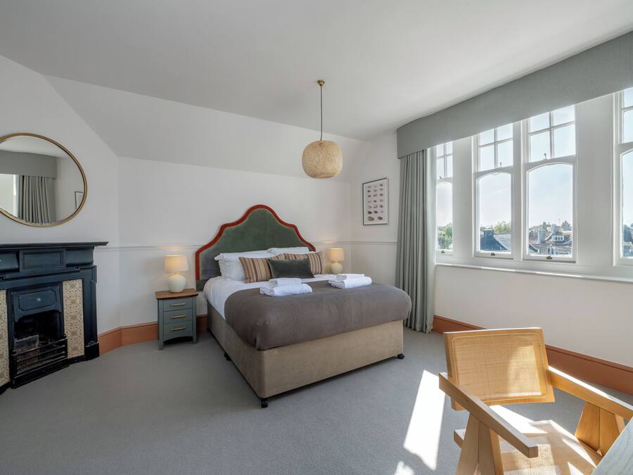 A neatly arranged bedroom with a bed, a small table with a lamp, a chair, a fireplace, and large windows offering a city view.