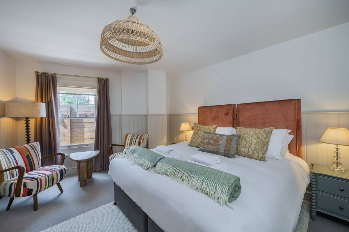 A hotel room with a large bed, two bedside tables, a chair, a small round table, a woven ceiling light, and a window with curtains.
