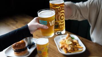 A person holding two glasses of beer, one with a logo and one with a design, on a table with food plates.