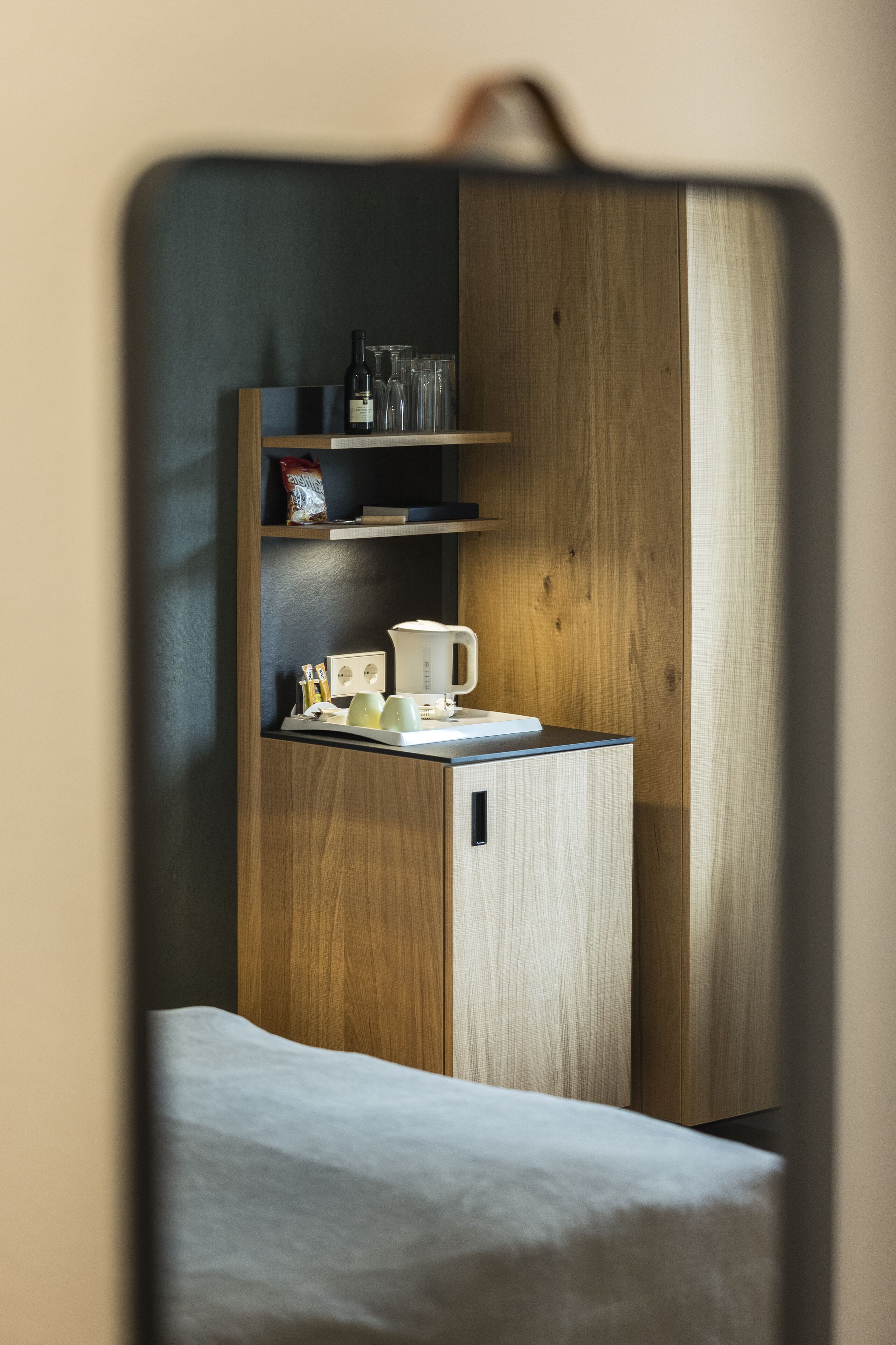 A compact bedroom with a wooden wardrobe, a small kitchenette with a coffee maker, and a bedside table.