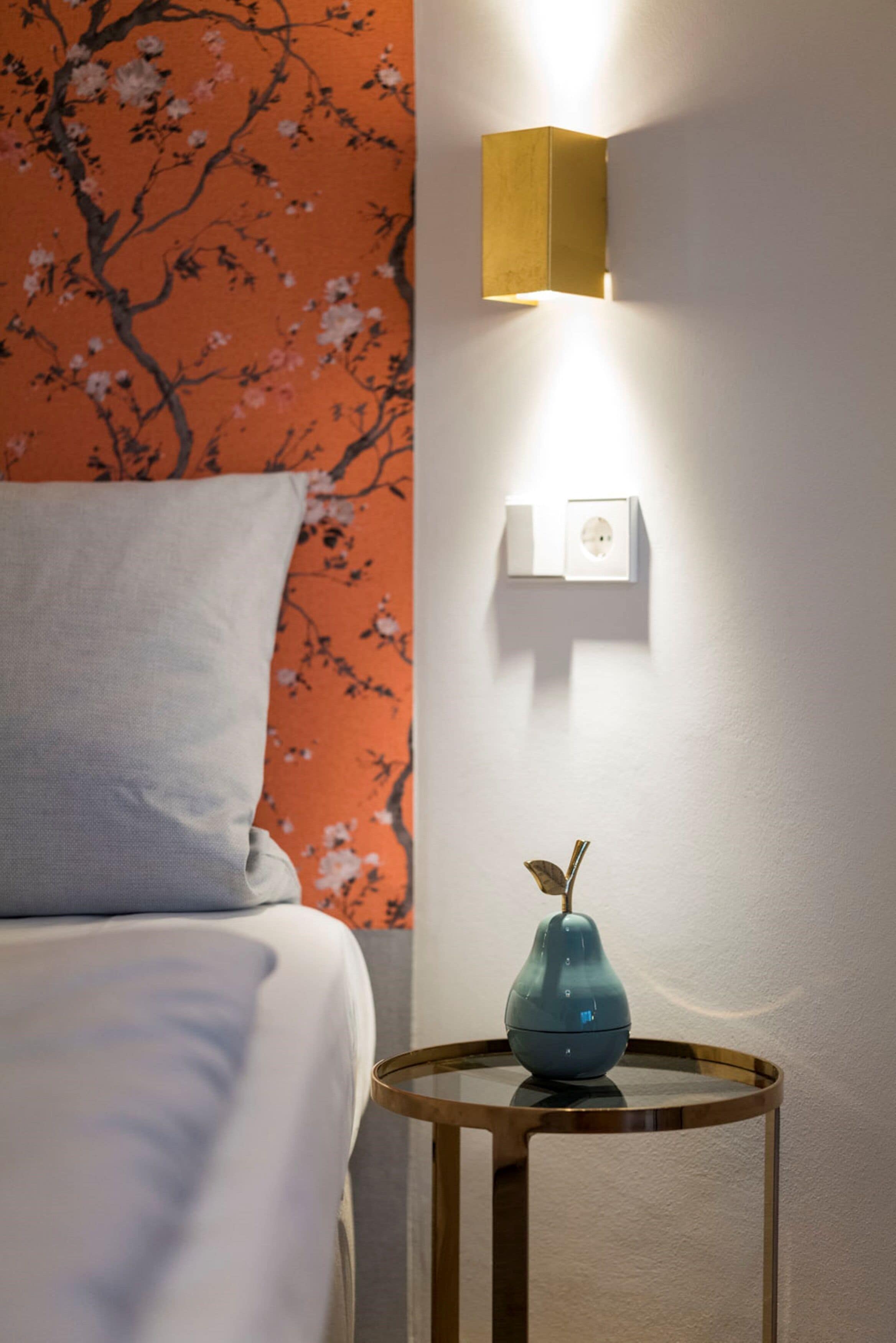 Habitación de hotel con cama, mesita de noche con un florero, lámpara de pared y toma de corriente.