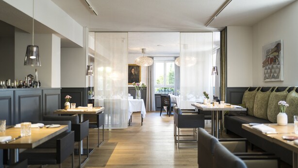 A modern dining area with wooden tables and chairs, a glass partition, and a view of the dining room in the background.