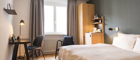 Habitación de hotel con cama, escritorio con silla, radiador, ventana con cortinas, armario de madera y estante de pared con objetos.