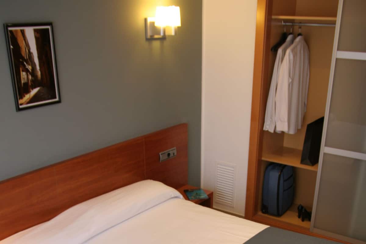 A hotel room with a large bed, a wardrobe, a bedside table, and a wall-mounted lamp.