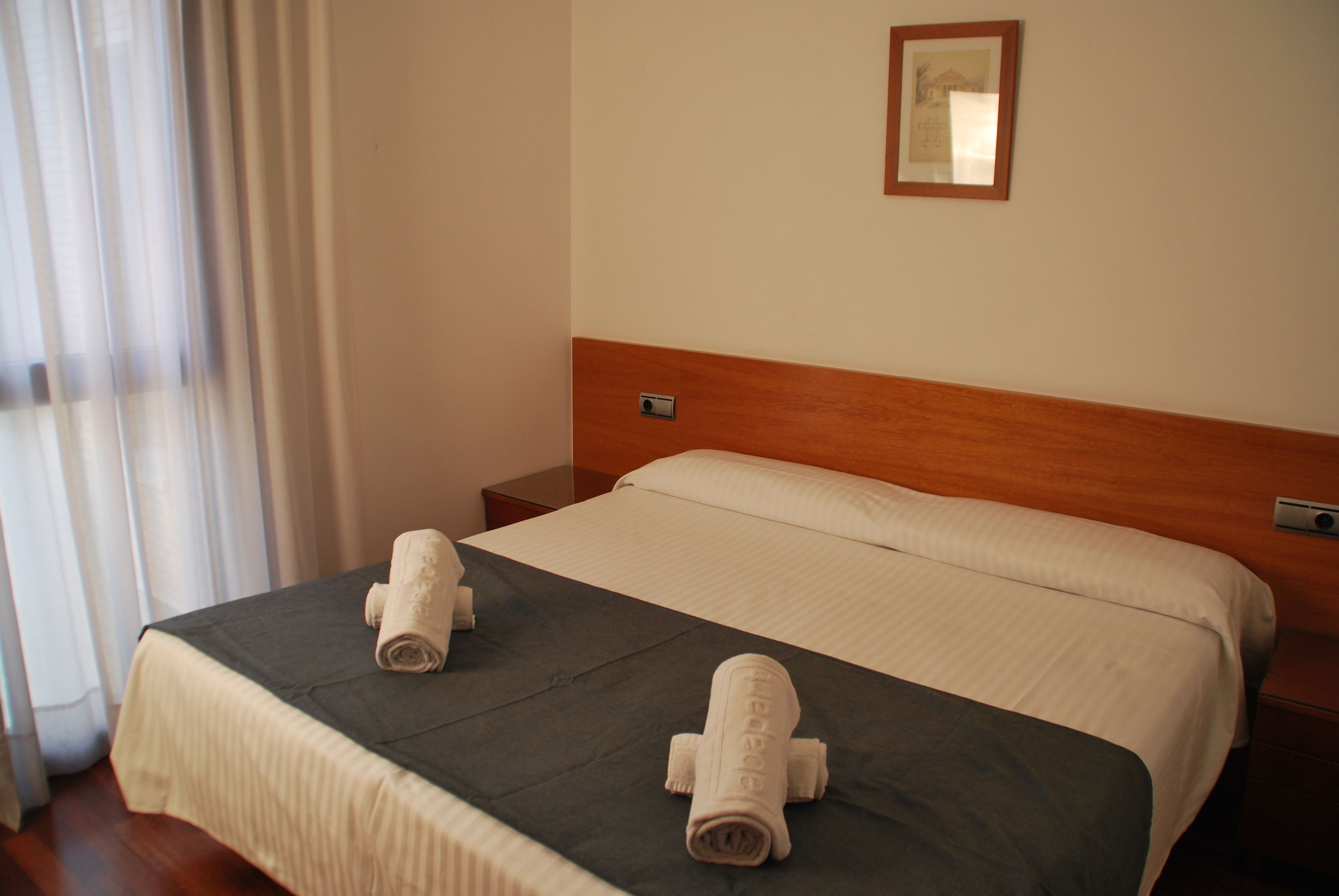 A hotel room with a bed, a nightstand, a wall-mounted mirror, and a window with curtains.