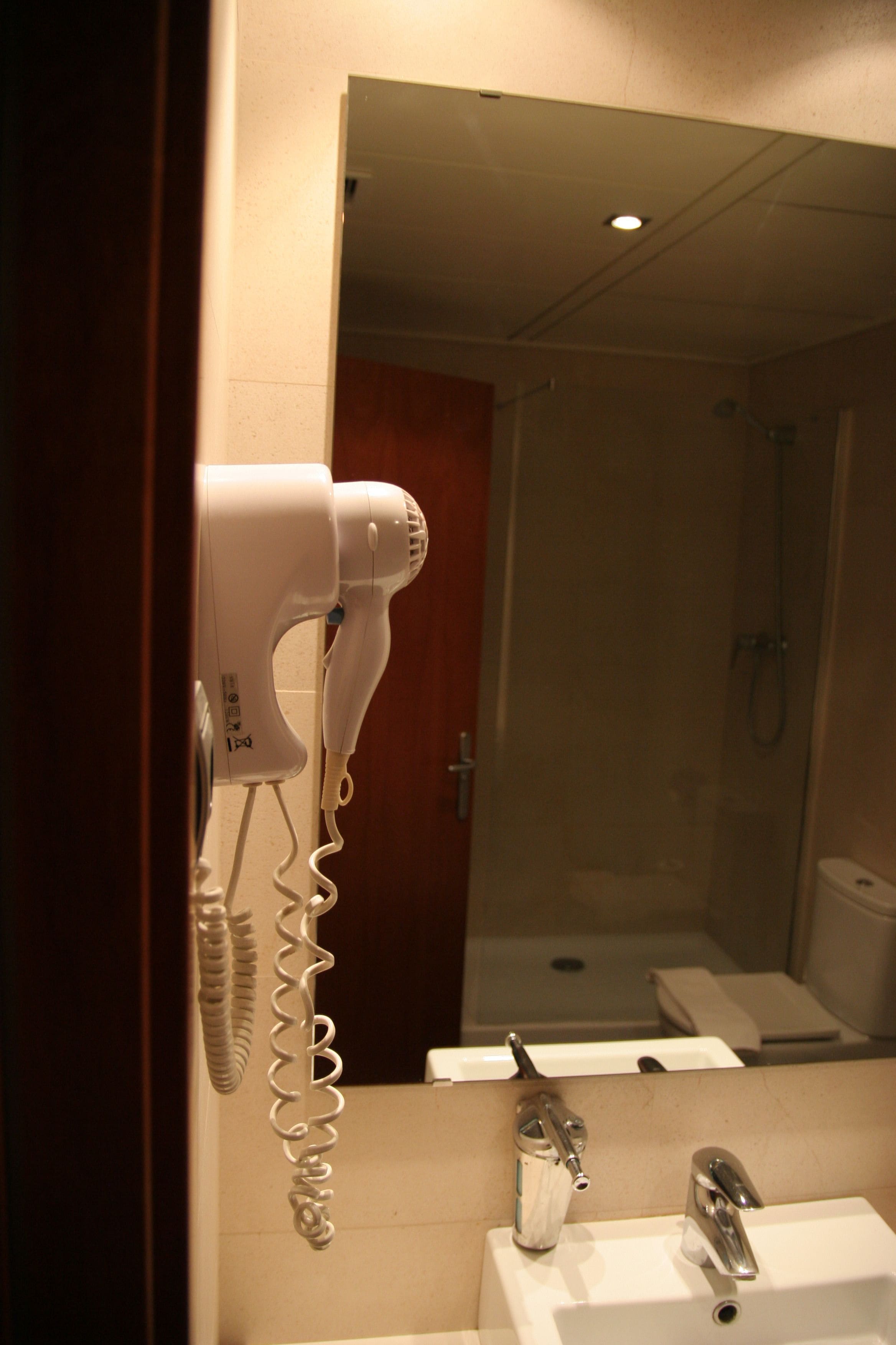 Un baño con un secador de pelo sujeto a la pared, un lavabo y un espejo que refleja una zona de ducha.
