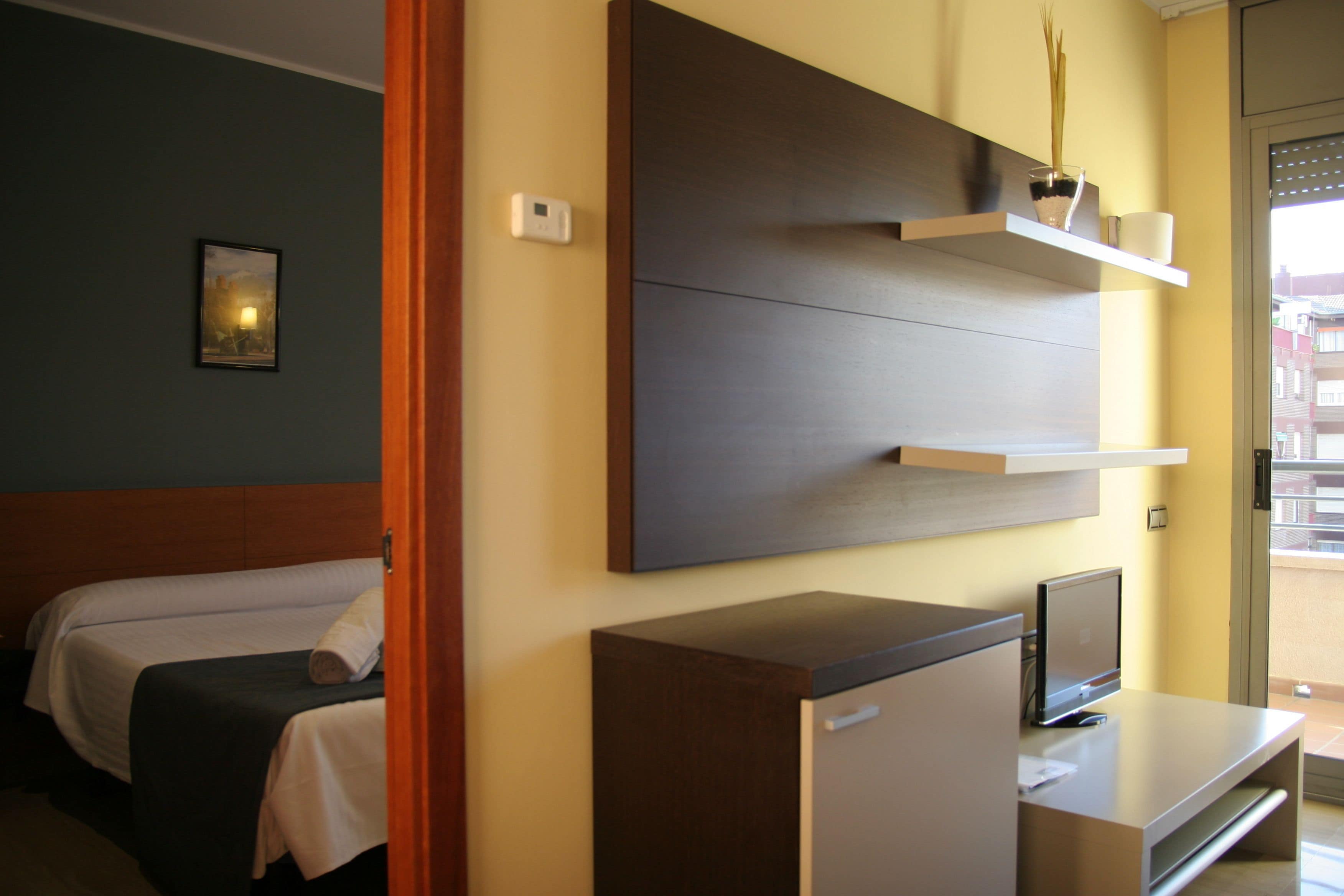 Une chambre d’hôtel avec un lit, un bureau avec un ordinateur, une étagère avec un vase et un tableau au mur.