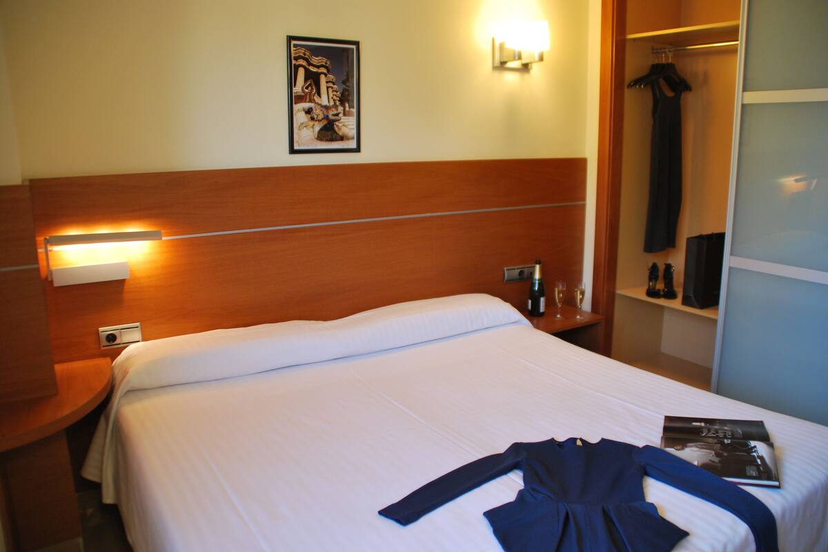 Ein Hotelzimmer mit einem ordentlich bezogenen Bett, einem Nachttisch mit Lampe, einem gerahmten Bild an der Wand und einem Schrank mit aufgehängter Kleidung.