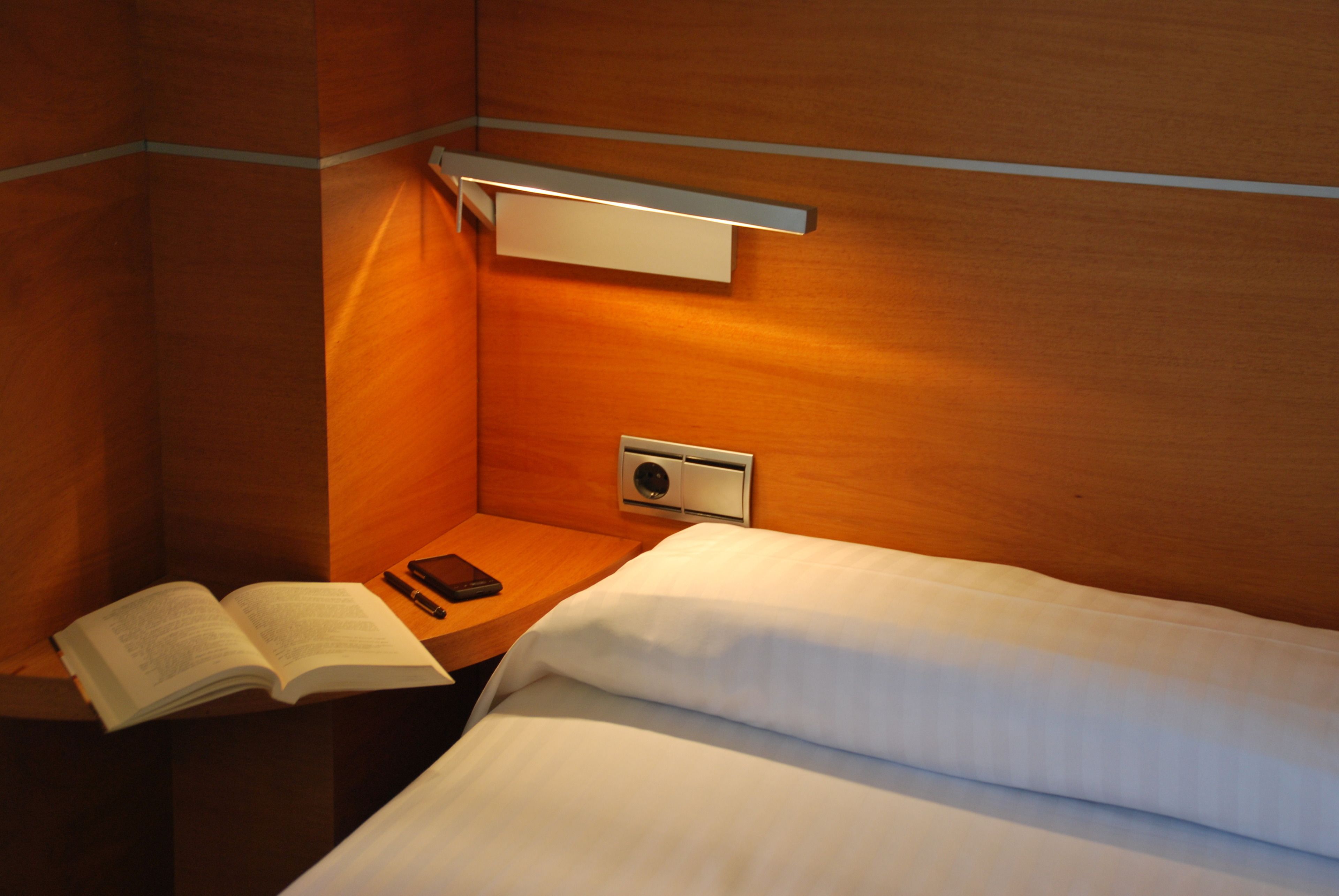 Une chambre d’hôtel avec un lit, une table de chevet sur laquelle repose un livre ouvert, une télécommande et un luminaire fixé au mur.