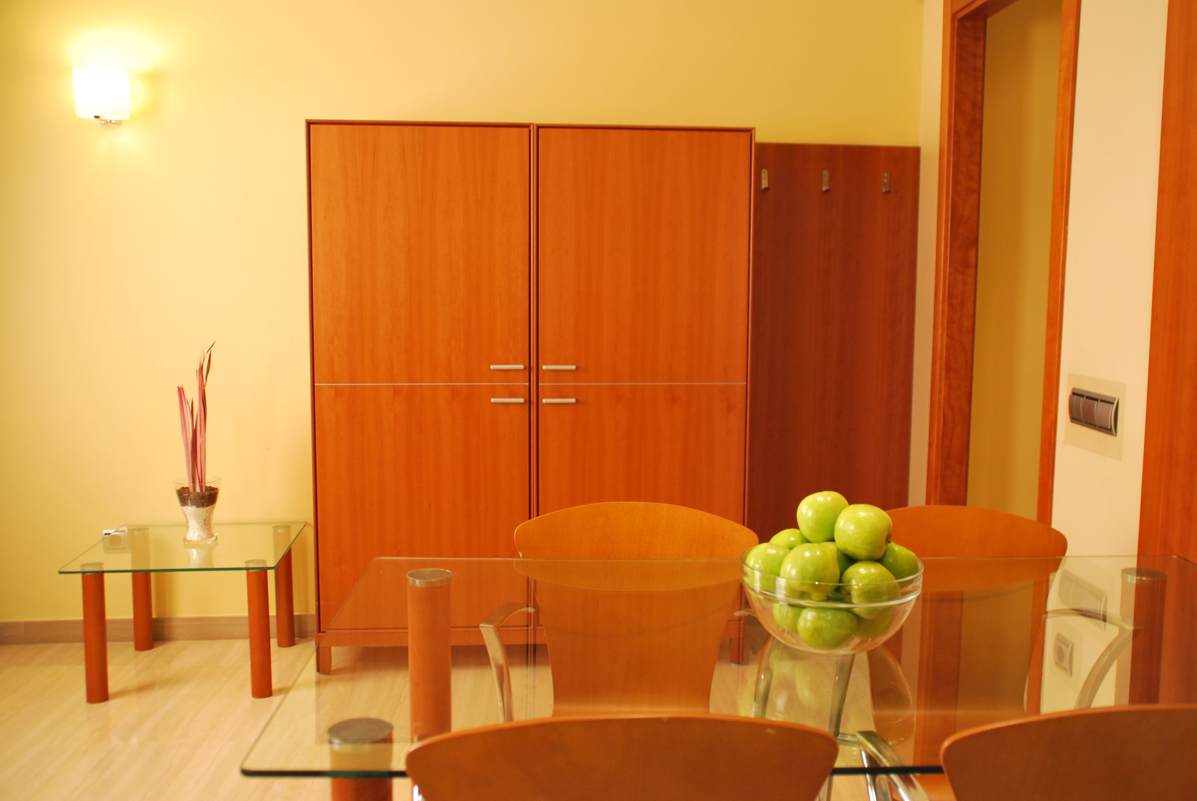 Un comedor moderno con una mesa de vidrio, dos sillas anaranjadas, un armario de madera y un bol con manzanas verdes.