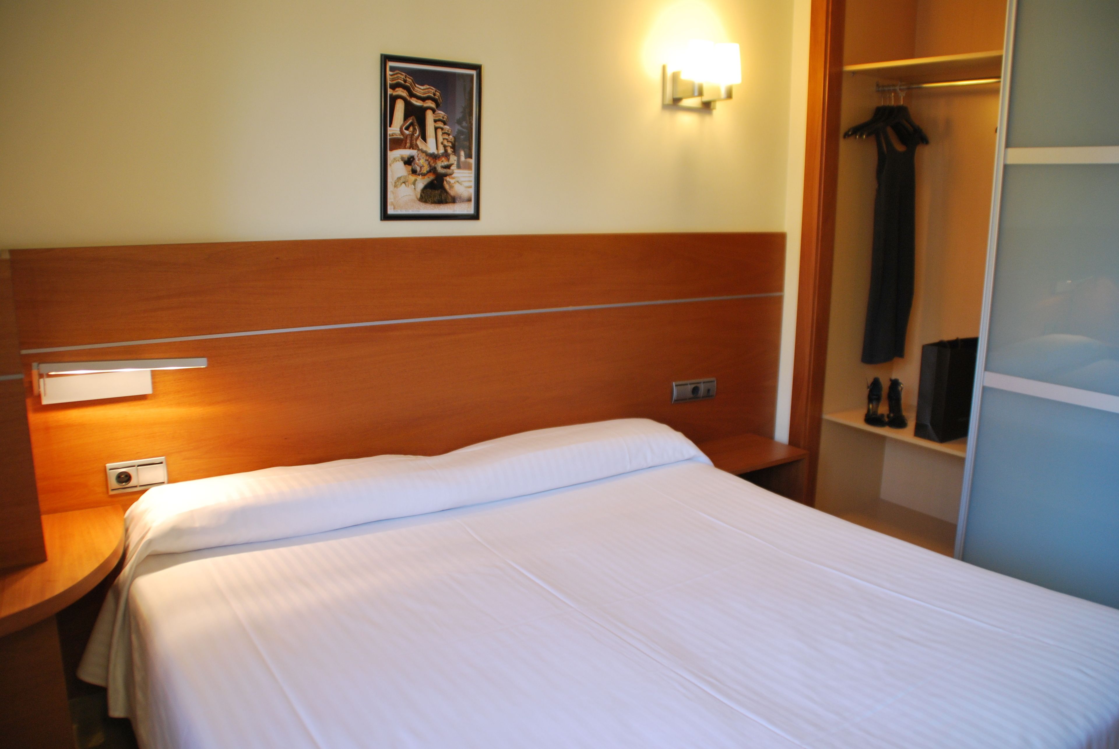 Una camera d'albergo con una testiera in legno, un copriletto bianco, un comodino, una lampada a parete, un quadro incorniciato e un armadio con un capo appeso.