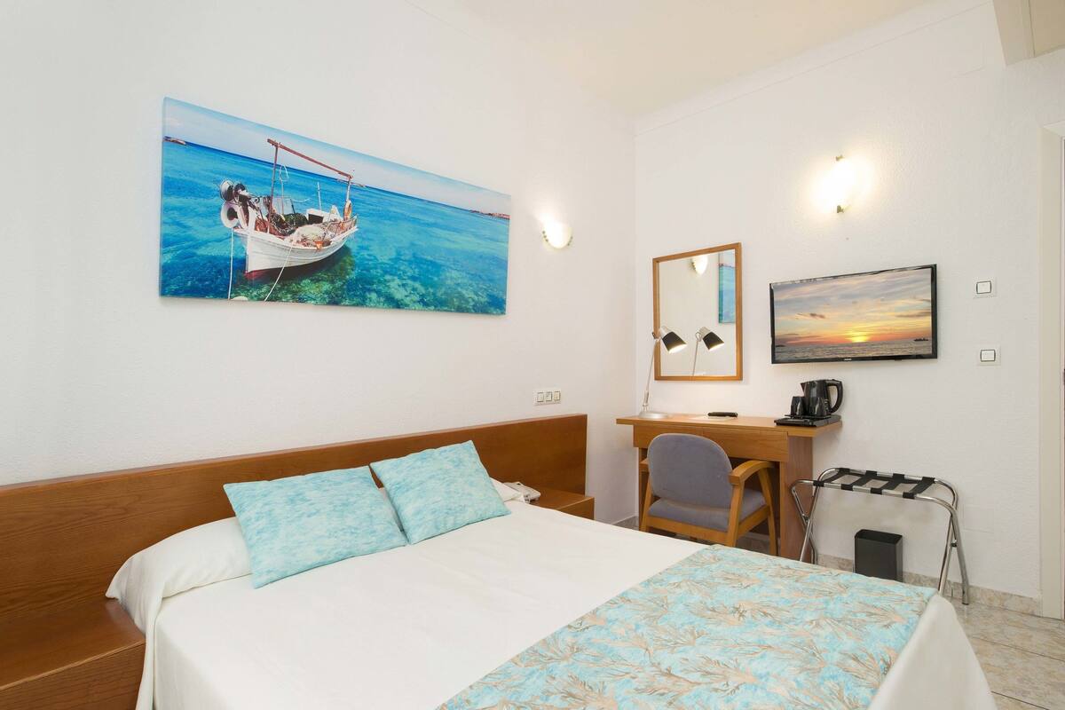 Quarto de hotel com cama, escrivaninha, cadeira, mesinha, espelho e um quadro de um barco na parede.
