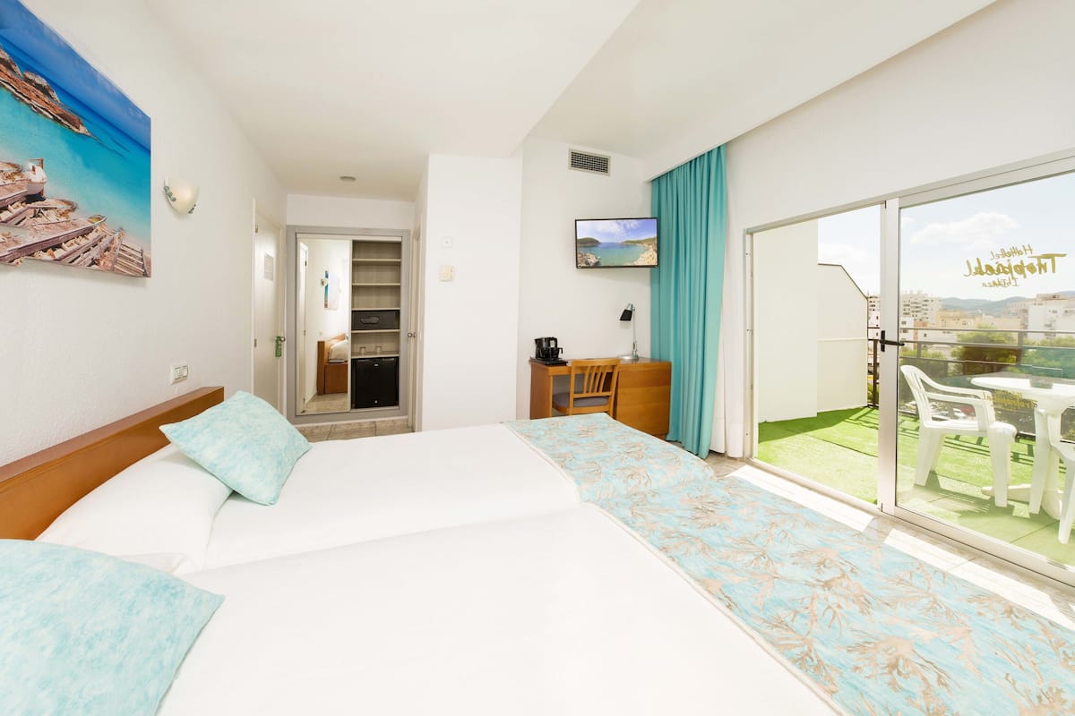 Quarto de hotel com cama, escrivaninha, televisão e varanda com mobiliário de exterior.