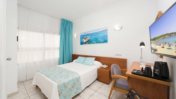 A hotel room with a bed, a desk, a chair, a TV, and a painting of a beach scene. - Hotel Tropical (Sant Antoni de Portmany)