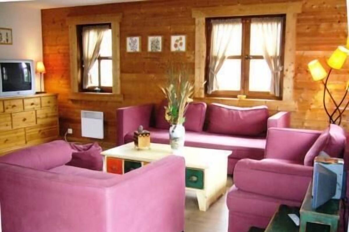 A cozy room with a wooden wall, a pink sofa set, a small table with a vase, and a TV on a wooden cabinet.