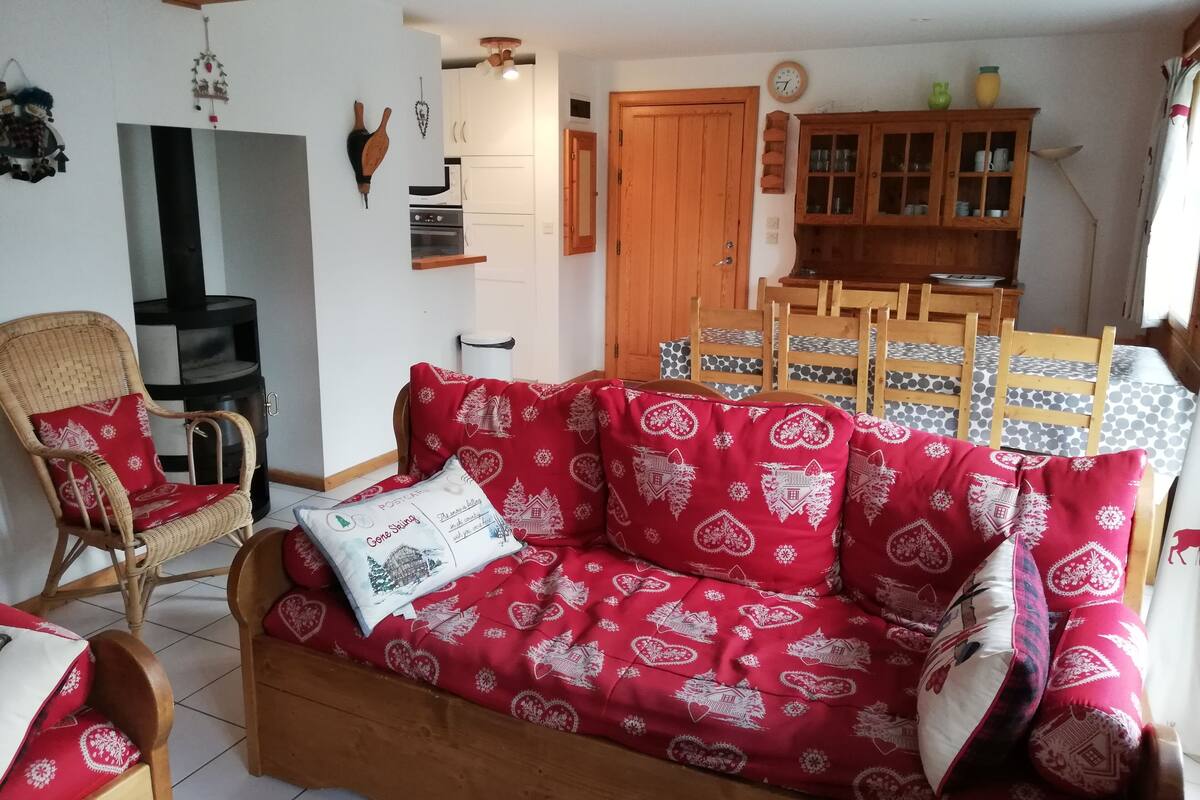 A cozy living room with a red patterned sofa, a wicker chair, a wooden dining table with chairs, and a kitchen area with cabinets and a stove.