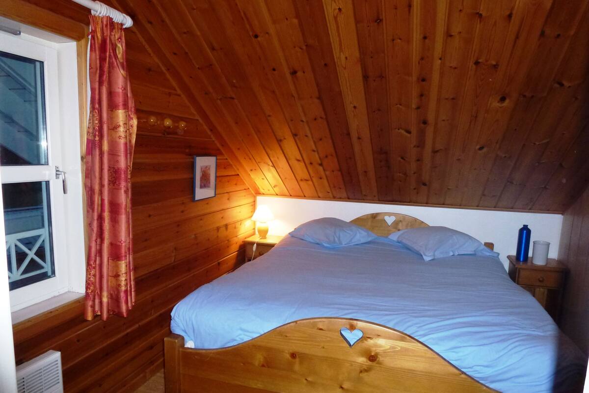 A wooden-paneled room with a bed, a window with curtains, a bedside lamp, and a small table with a bottle and cup.