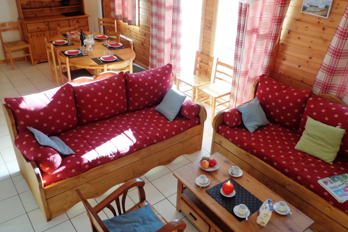 A cozy cabin interior with a wooden dining table set for four, two red sofas with star patterns, and a small wooden table with cups and a candle.