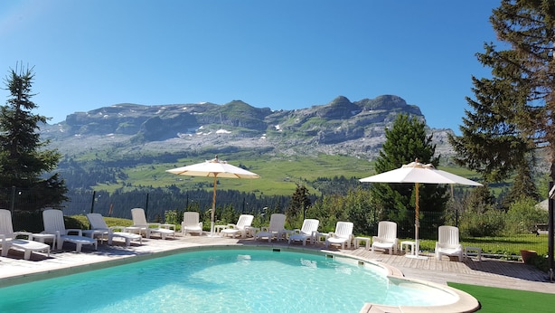 Een zwembad met witte ligbedden en parasols, omgeven door een houten terras, met uitzicht op een berg in de verte.